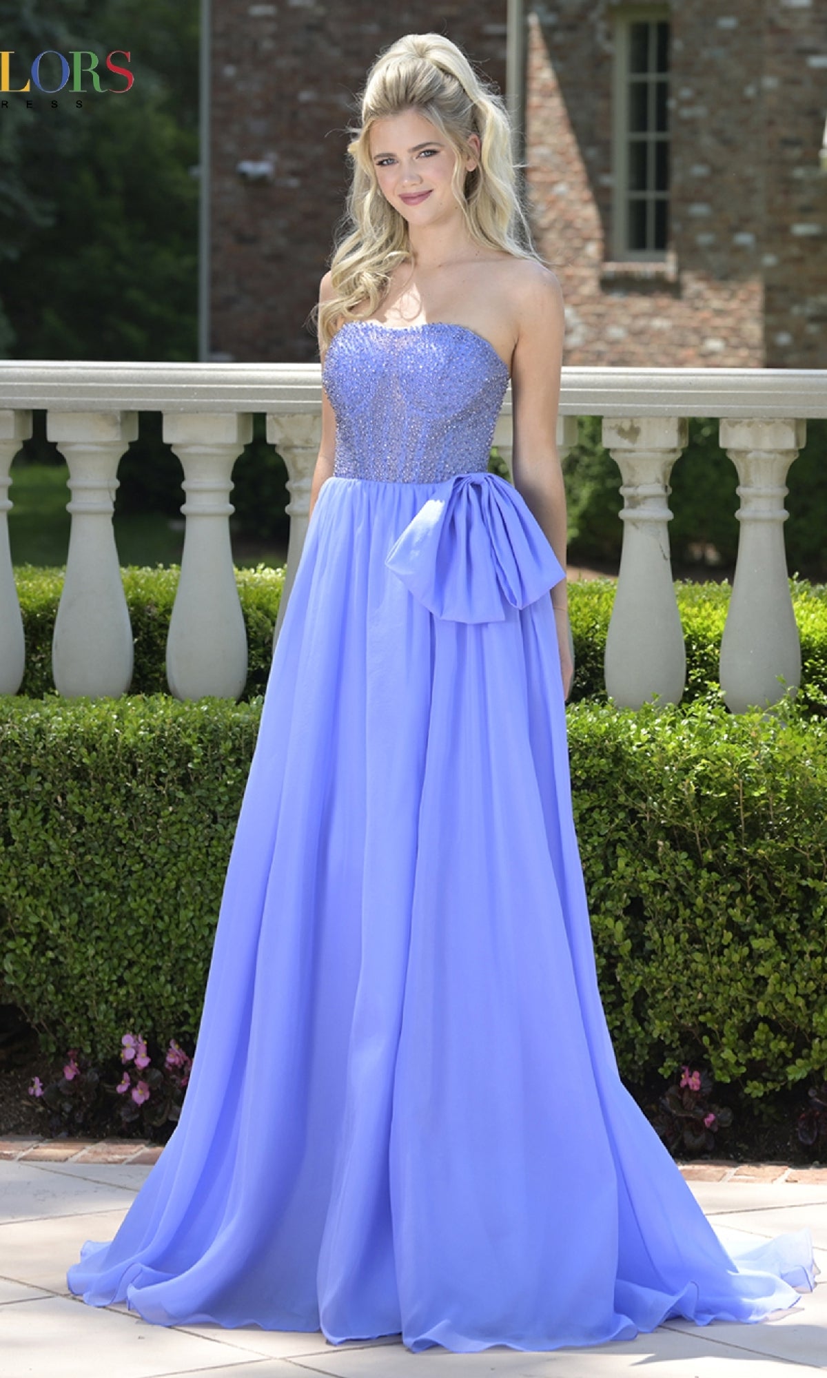 A woman stands outdoors wearing the Colors Dress 3843, a long lavender prom dress with a strapless textured bodice, large waist bow, and flowing chiffon A-line skirt.